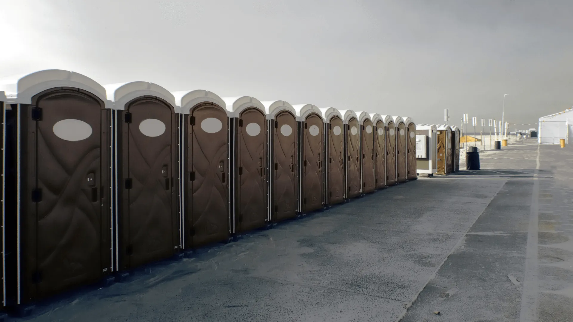 A long row of portable toilets on a concrete surface under a gray sky, with a deserted street in the background.